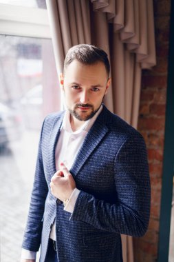 Takım elbiseli genç bir adam ağır perdeli bir pencerenin yanında duruyor. Pencerenin önünde penceresinde antika perdeleri olan bir adam.