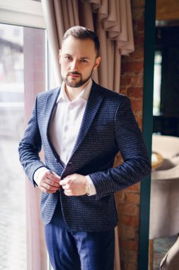 Takım elbiseli genç bir adam ağır perdeli bir pencerenin yanında duruyor. Pencerenin önünde penceresinde antika perdeleri olan bir adam.