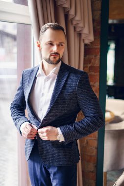 Takım elbiseli genç bir adam ağır perdeli bir pencerenin yanında duruyor. Pencerenin önünde penceresinde antika perdeleri olan bir adam.