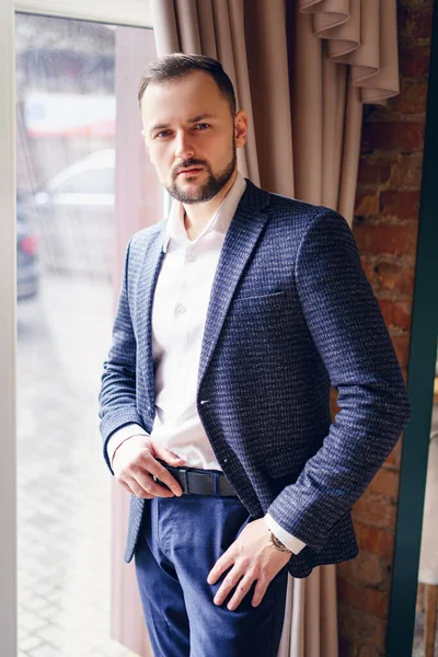 A young man in a business suit stands near a window with heavy curtains. A man at the window in a room with vintage curtains at the window.