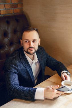 Düzgün sakallı genç bir adam, iş elbisesi giymiş, akıllı bir telefon ve kahve ile masada bir fincan kahve ile iş adamı oturarak telefon, çay ya da restoranda kahve içmeyi düşünüyor.