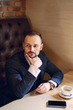 Düzgün sakallı genç bir adam, iş elbisesi giymiş, akıllı bir telefon ve kahve ile masada bir fincan kahve ile iş adamı oturarak telefon, çay ya da restoranda kahve içmeyi düşünüyor.