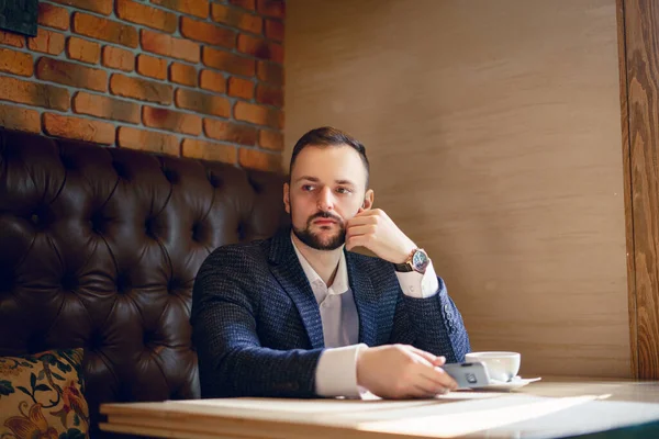 A young man in a business suit is sitting at a table in a restaurant with a cup of coffee. A businessman drinking coffee from a white cup in a restaurant or cafe.
