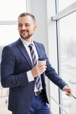 A young man with a neat little beard is dressed in a business suit with a white shirt and tie with a disposable paper cup of tea or coffee by the large window. Positive young businessman at the window