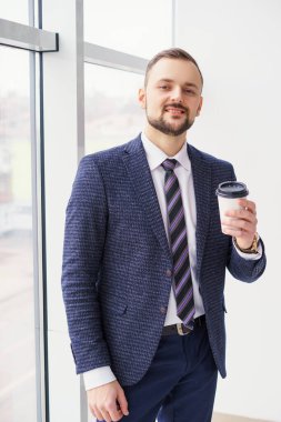A young man with a neat little beard is dressed in a business suit with a white shirt and tie with a disposable paper cup of tea or coffee by the large window. Positive young businessman at the window