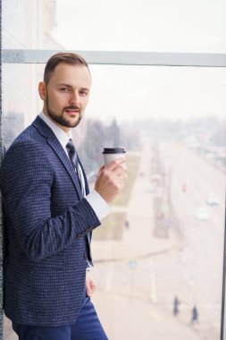A young man with a neat little beard is dressed in a business suit with a white shirt and tie with a disposable paper cup of tea or coffee by the large window. Positive young businessman at the window