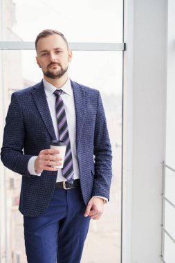 A young man with a neat little beard is dressed in a business suit with a white shirt and tie with a disposable paper cup of tea or coffee by the large window. Positive young businessman at the window