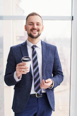 A young man with a neat little beard is dressed in a business suit with a white shirt and tie with a disposable paper cup of tea or coffee by the large window. Positive young businessman at the window