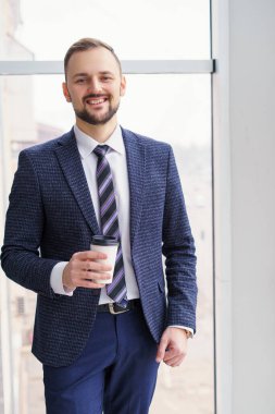 A young man with a neat little beard is dressed in a business suit with a white shirt and tie with a disposable paper cup of tea or coffee by the large window. Positive young businessman at the window