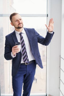 A young man with a neat little beard is dressed in a business suit with a white shirt and tie with a disposable paper cup of tea or coffee by the large window. Positive young businessman at the window