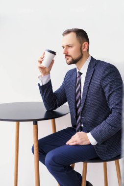 A young man in a business suit at a small round table drinking coffee or tea from a white cup. A young businessman is relaxing over a cup of coffee.