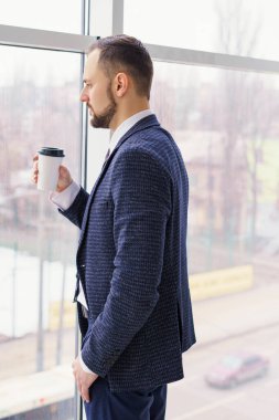 A young man in a business suit with a cup of coffee stands at a large window and looks out.