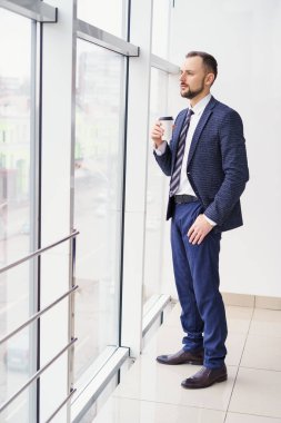 A young man in a business suit with a cup of coffee stands at a large window and looks out.