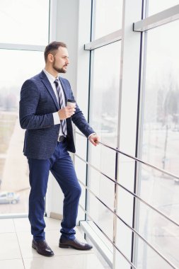 A young man in a business suit with a cup of coffee stands at a large window and looks out.