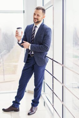 A young man in a business suit with a cup of coffee stands at a large window and looks out. Young businessman on the break
