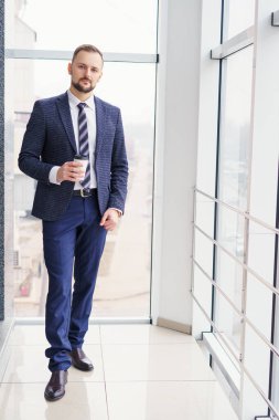 A young man in a business suit with a cup of coffee stands at a large window and looks out. Young businessman on the break