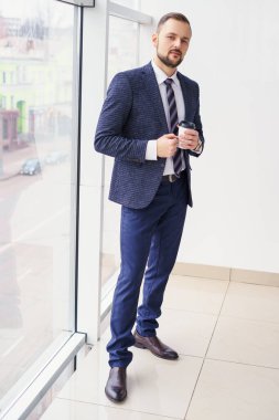A young man in a business suit with a cup of coffee stands at a large window and looks out. Young businessman on the break