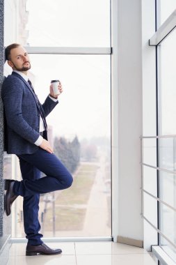A young man in a business suit with a cup of coffee stands at a large window and looks out. Young businessman on the break