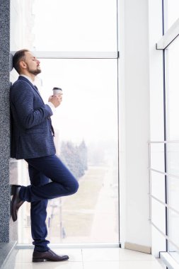 A young man in a business suit with a cup of coffee stands at a large window and looks out. Young businessman on the break