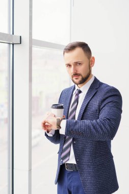 A young man in a business suit looks at his watch with a cup of coffee. A businessman in a shirt and tie and a dark jacket. A bearded man in a cafe or restaurant.
