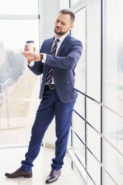 A young man in a business suit looks at his watch with a cup of coffee. A businessman in a shirt and tie and a dark jacket. A bearded man in a cafe or restaurant.