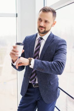 A young man in a business suit looks at his watch with a cup of coffee. A businessman in a shirt and tie and a dark jacket. A bearded man in a cafe or restaurant.