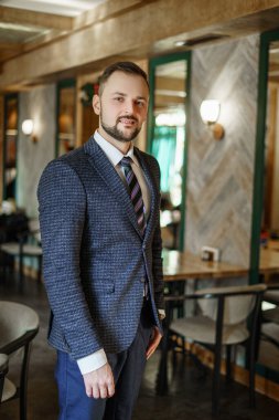 A young man in a business suit looks away. A businessman in a shirt and tie and a dark jacket. A bearded man in a cafe or restaurant.