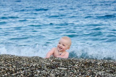 A little boy is lying on the beach playing with pebbles. A small child on the beach of pebbles. In the background is a blue sea out of focus and with waves.