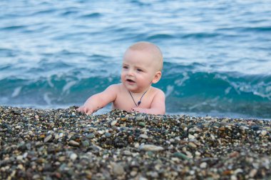 A little boy is lying on the beach playing with pebbles. A small child on the beach of pebbles. In the background is a blue sea out of focus and with waves.