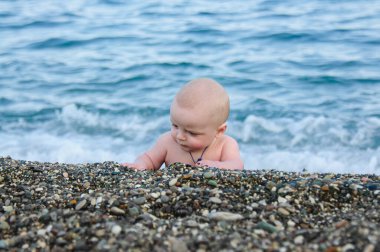 A little boy is lying on the beach playing with pebbles. A small child on the beach of pebbles. In the background is a blue sea out of focus and with waves.