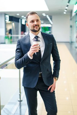 A young bearded man dressed in a business suit at the mall with a cup of coffee. A young man in an elegant business suit strolls through a mall drinking coffee as he goes