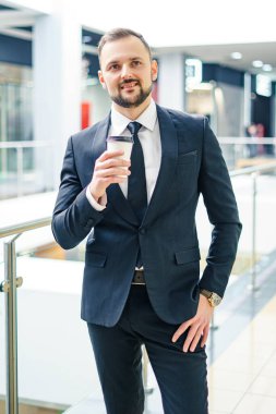 A young bearded man dressed in a business suit at the mall with a cup of coffee. A young man in an elegant business suit strolls through a mall drinking coffee as he goes