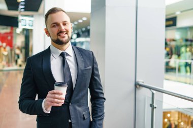A young bearded man dressed in a business suit at the mall with a cup of coffee. A young man in an elegant business suit strolls through a mall drinking coffee as he goes