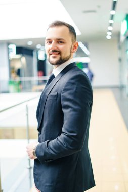 A young man with a beard dressed in a classic business suit. Young businessman in a mall wearing a black suit and black tie