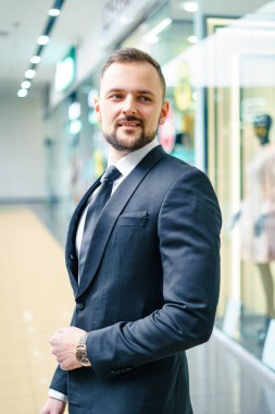 A young man with a beard dressed in a classic business suit. Young businessman in a mall wearing a black suit and black tie