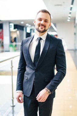 A young man with a beard dressed in a classic business suit. Young businessman in a mall wearing a black suit and black tie