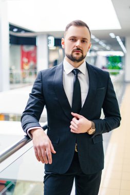 A young man with a beard dressed in a classic business suit. Young businessman in a mall wearing a black suit and black tie