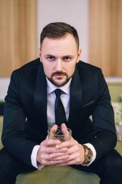 A young man with a beard, dressed in a classic business suit, sitting in the hands of a glass of whiskey. Portrait of a man relaxing with a glass of hard liquor.