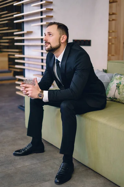 A young man with a beard, dressed in a classic business suit, sitting in the hands of a glass of whiskey. Portrait of a man relaxing with a glass of hard liquor.