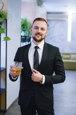 Sakallı, takım elbiseli, elinde bir bardak viski tutan genç bir adam. Restoranda alkol içen bir işadamı.