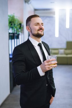 Sakallı, takım elbiseli, elinde bir bardak viski tutan genç bir adam. Restoranda alkol içen bir işadamı.