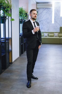 Sakallı, takım elbiseli, elinde bir bardak viski tutan genç bir adam. Restoranda alkol içen bir işadamı.