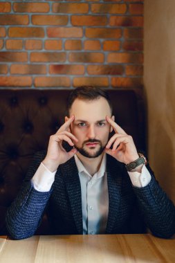 Küçük sakallı genç bir adam ellerini şakağına dayamış bir masada oturuyor. Restoranda ya da kafede beyaz gömlekli ve kravatlı bir adam..