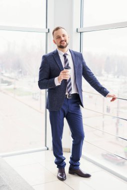 A young bearded man dressed in a business suit at the mall with a cup of coffee. A young man in an elegant business suit strolls through a mall drinking coffee as he goes
