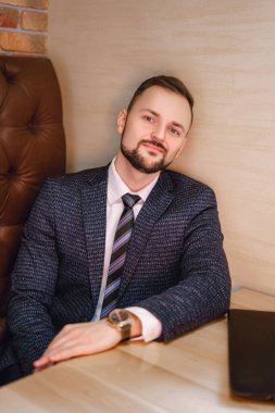 Küçük sakallı genç bir adam bir masada oturur. Restoranda ya da kafede beyaz gömlekli ve kravatlı bir adam..