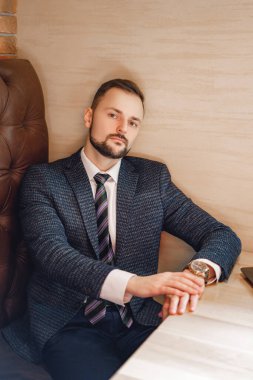 Küçük sakallı genç bir adam bir masada oturur. Restoranda ya da kafede beyaz gömlekli ve kravatlı bir adam..