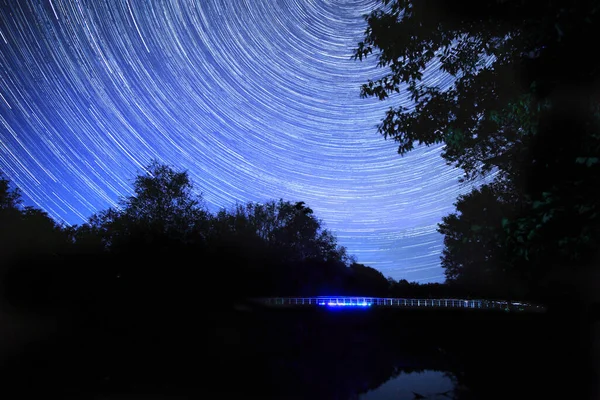 The night movement of stars, star tracks over the river and the bridge. The river banks are overgrown with trees.