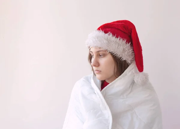 A sad tired girl in a Santa hat wrapped in a white blanket — Stock Photo, Image