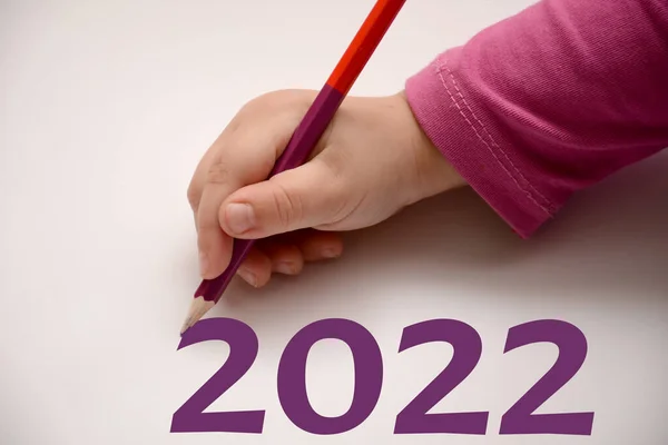 Een kind op een wit laken schrijft met een gekleurd potlood 2022. Het concept van de vakantie. Kinderen hand in een roze trui met een potlood. — Stockfoto
