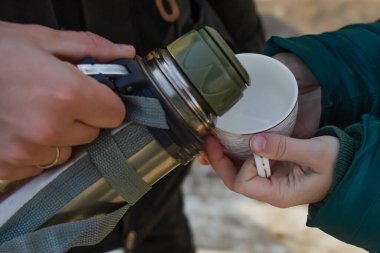Çelik termos tutan bir adamın eli başka bir kadının elindeki beyaz fincana kahve ya da çay dökmek istiyor..